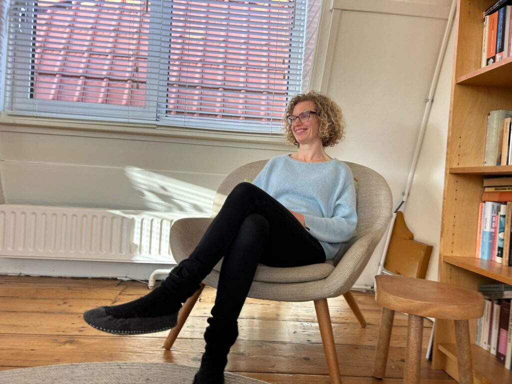 Eltica zit tijdens een gesprek in een grijze fauteuil naast de boekenkast.